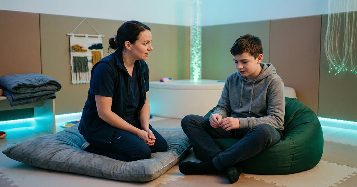 A care worker kneeling beside a teenager in a calming sensory room, demonstrating de-escalation and emotional regulation