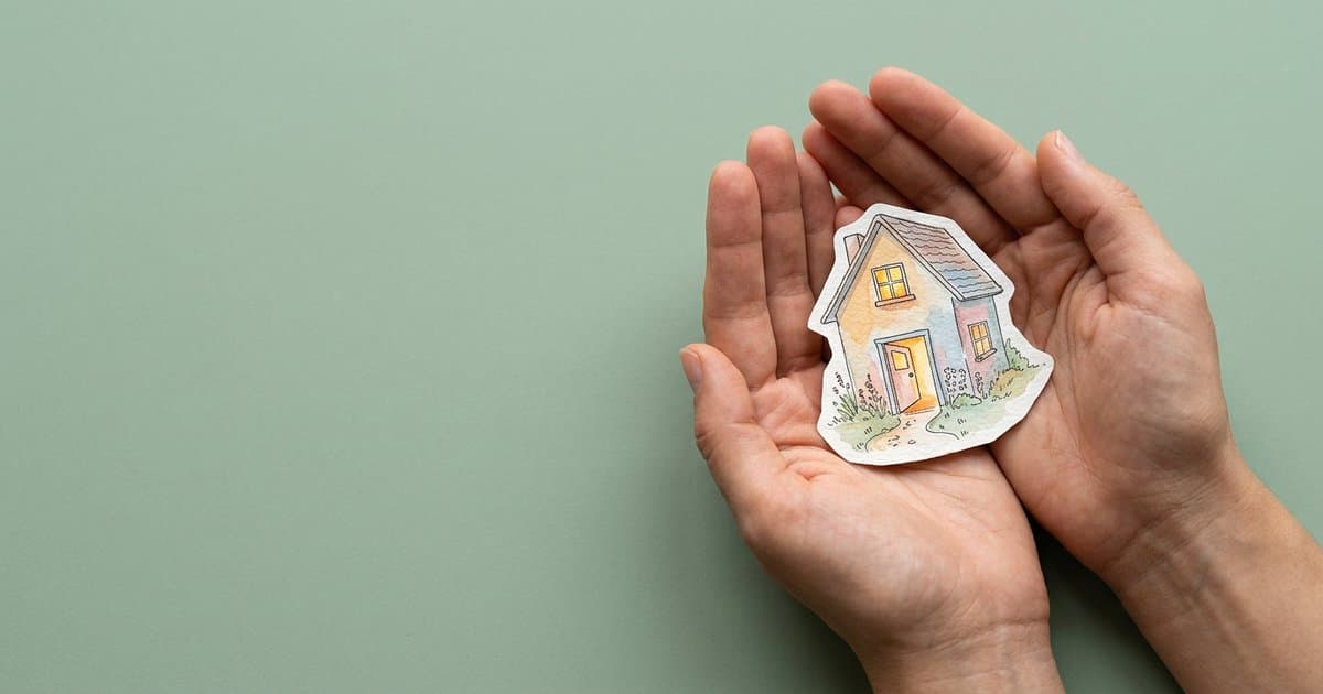 Hands holding a watercolour illustration of a small house with its front door open and warm light spilling onto the path